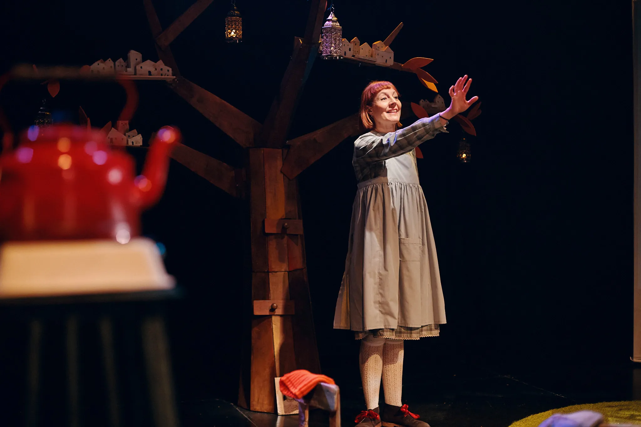 Ana Torres Lara, vestida de niña con abrigo de cuadros, en escena junto a un árbol con una tetera roja colgada