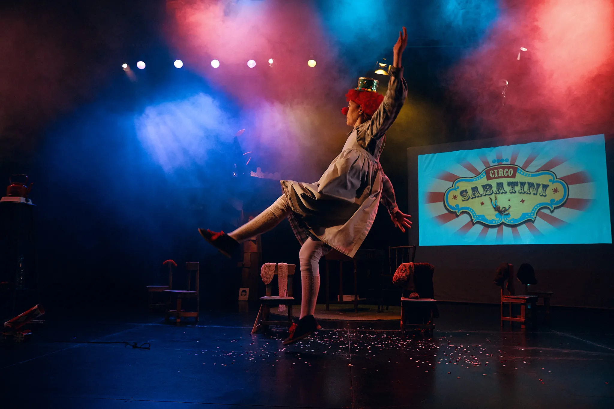 La actriz salta en escena bajo luces de colores ante una pantalla con un cartel de circo
