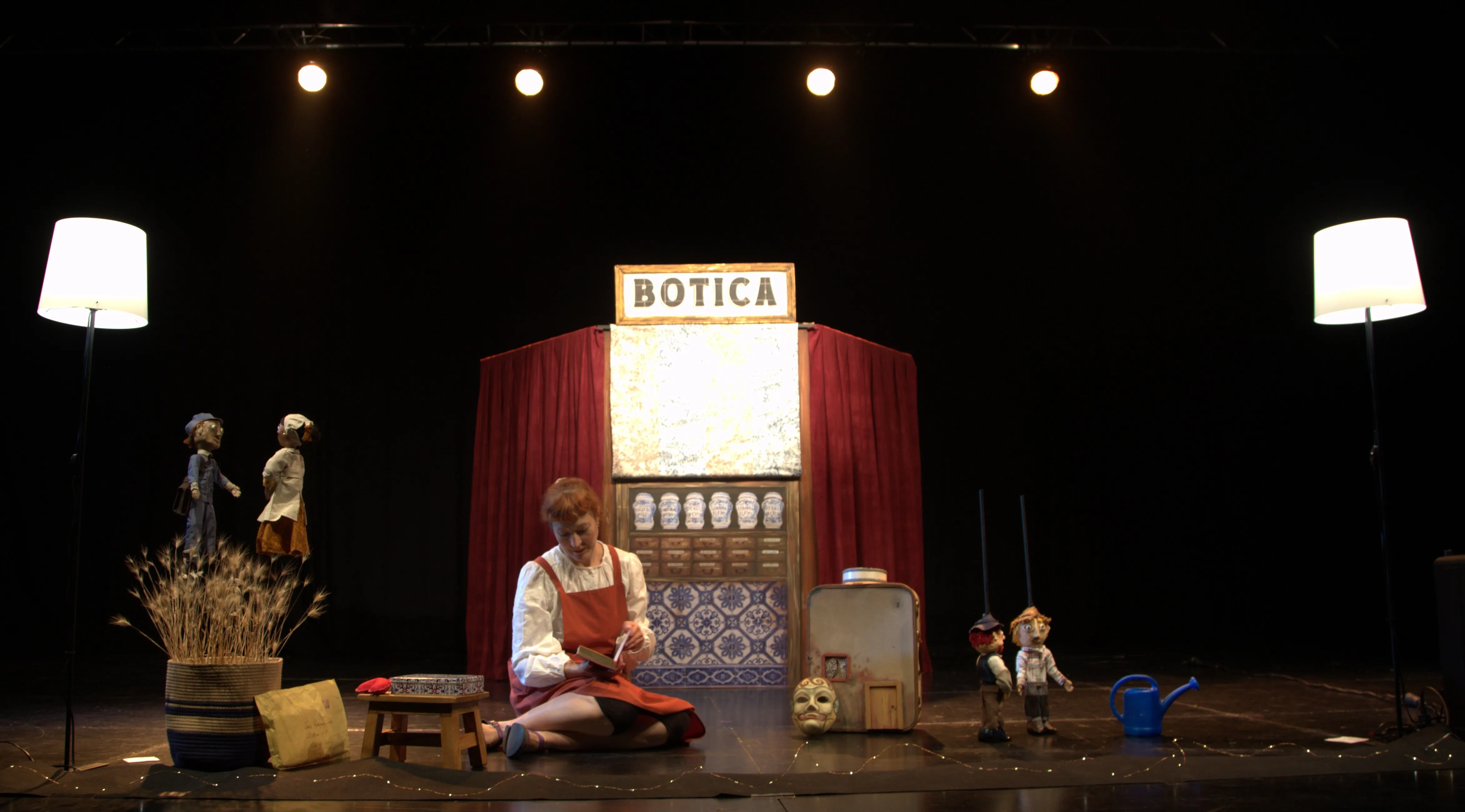 Ana Torres sentada en el escenario entre títeres, una maleta y el mostrador de la botica