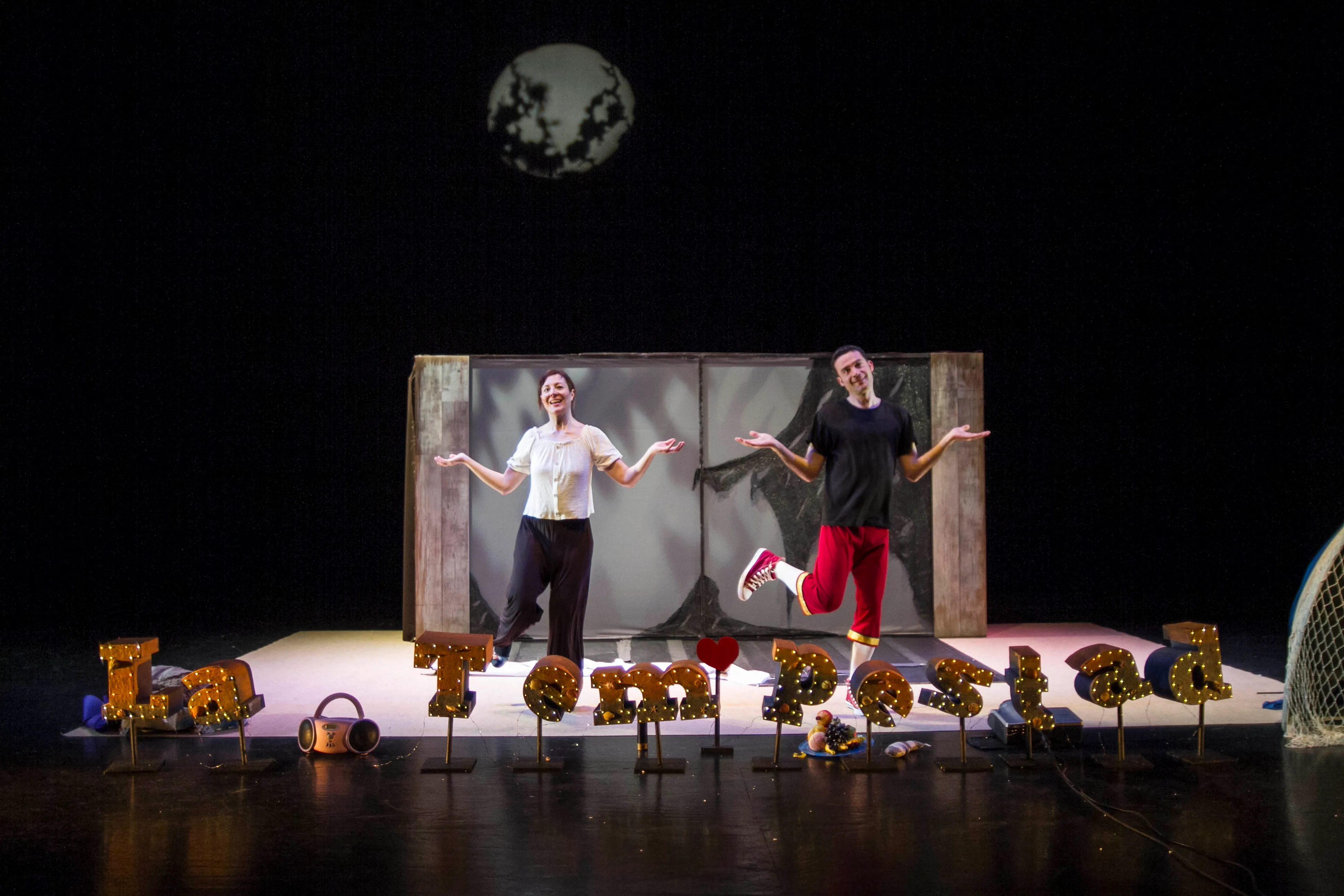 Los dos actores en escena bajo una luna, con el título La Tempestad escrito en letras de luz sobre el suelo