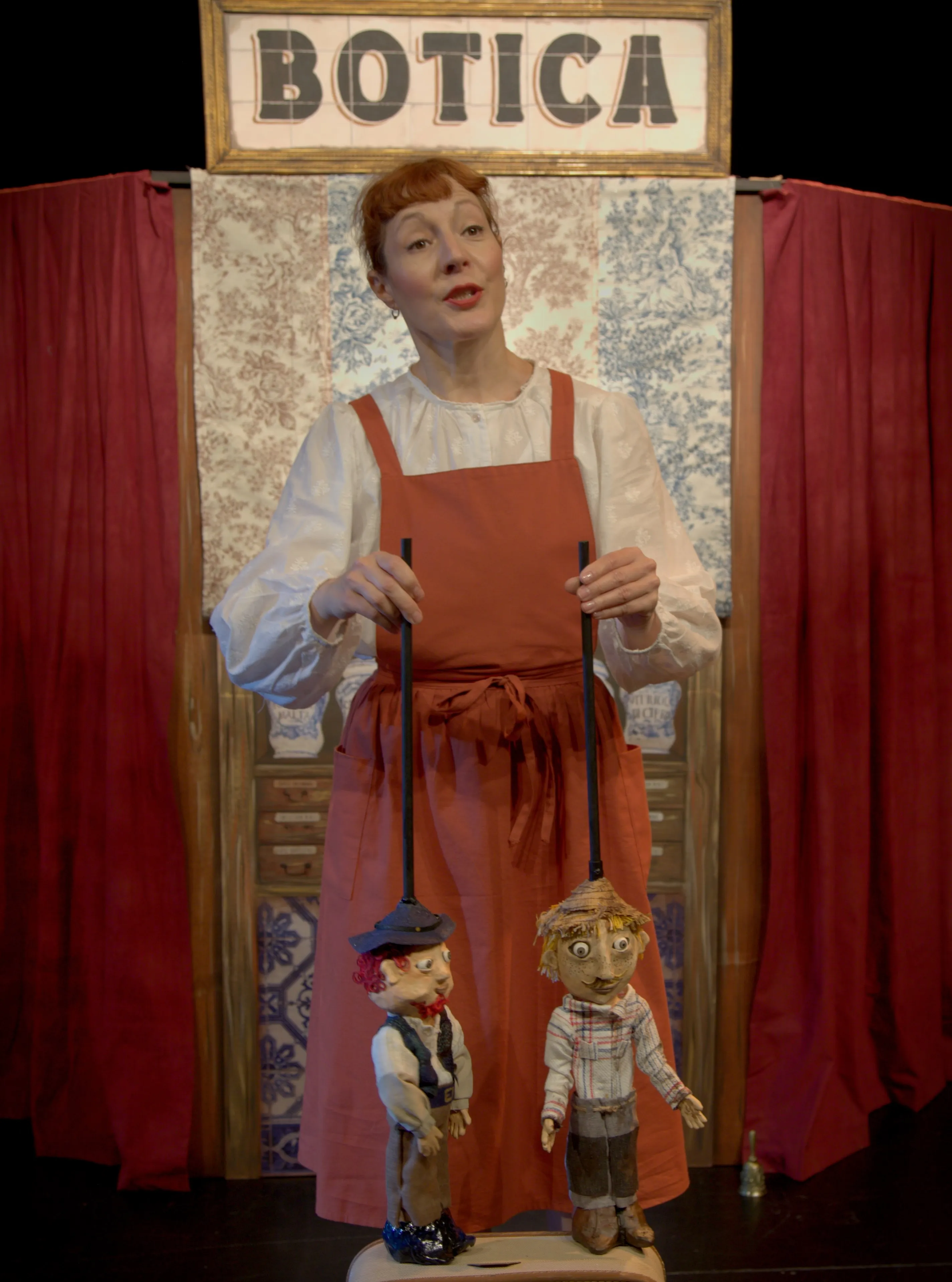 Jara, con delantal rojo, maneja dos marionetas delante del cartel de la botica